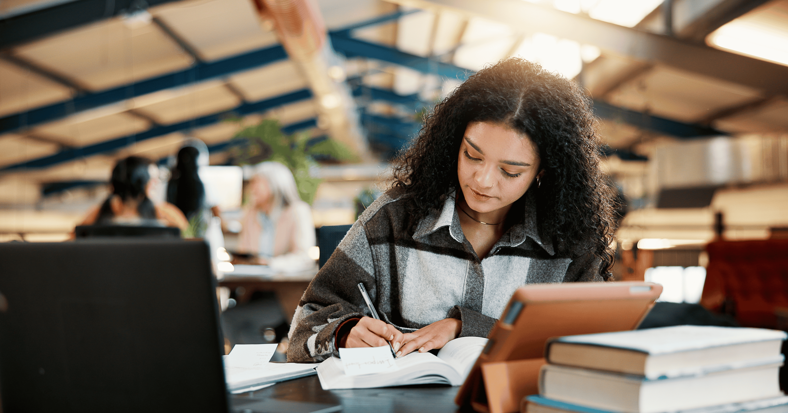 Woman studying by writing in a notebook in library with a laptop, tablet and books surrounding her for the Writing for Publication Network Cafe event